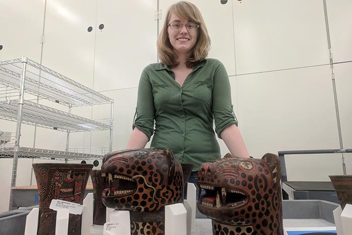 Allison Curley stands behind two wooden qeros in the shape of jaguar heads.