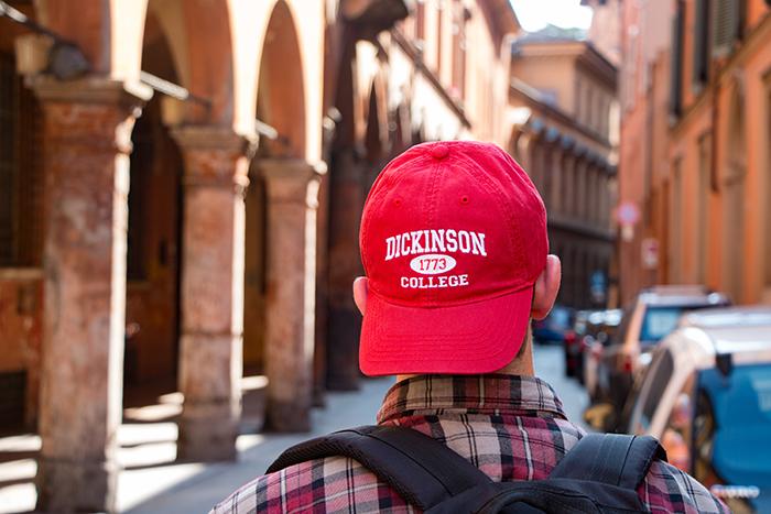Photo of a young man wearing a Dickinson College baseball cap, walking down a street in a European city.