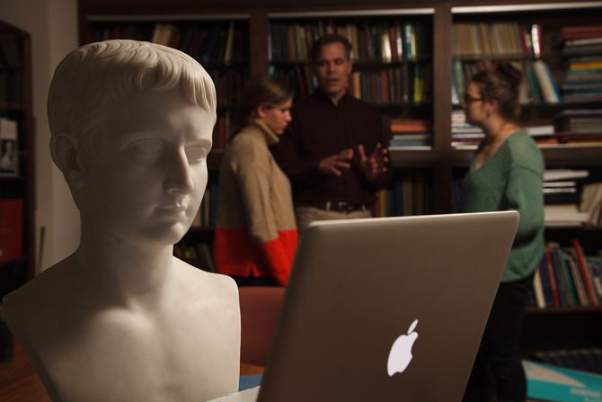 A classic bust looking at a computer screen in the foreground while a professor and two students talk in the background. 