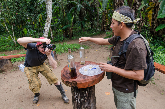 A man at right has placed three bottles with dark liquids on a table at center, while Andrew Bale crouches to photograph them at left.