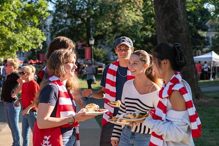 Several college students converse and enjoy cookout food on a leafy college campus.