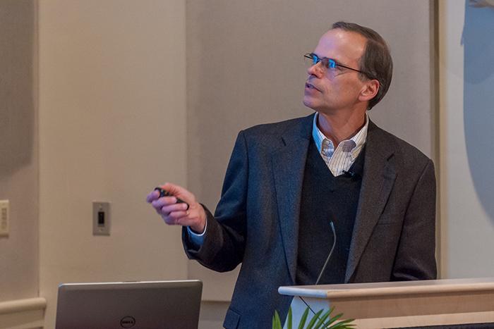 Photo of John Henson standing at a lectern, gesturing toward a screen showing a PowerPoint presentation.