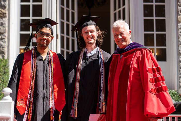 Three men in academic regalia stand in front of a limestone building.