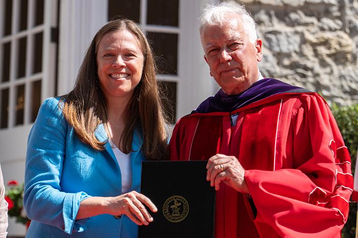 Photo of a woman in a blue blazer and a man in a red academic robe holding a folio in front of a limestone building.