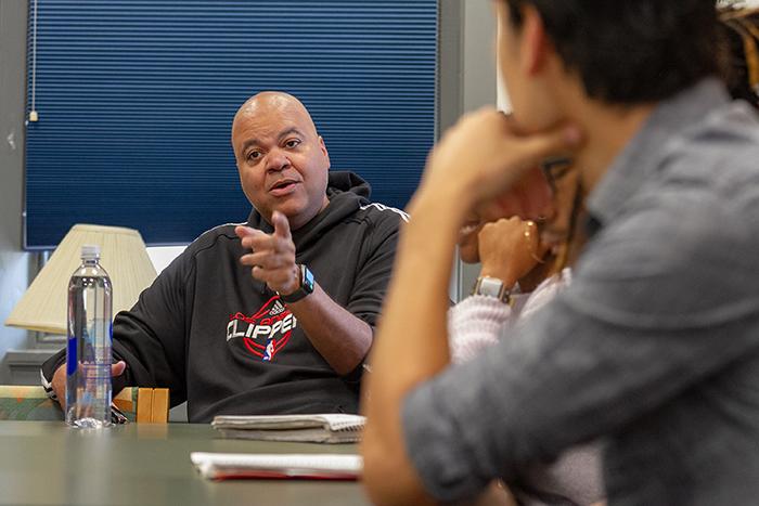 Ed Ricourt points his finger toward the camera, as two students lean on a table and listen to him speak.