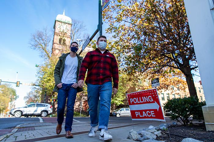 Two men walk on a sidewalk with a bell tower and trees behind them.