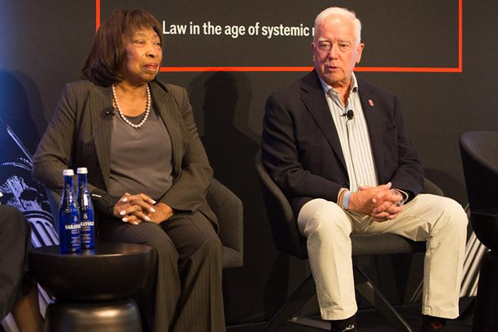 Two people, a man and a woman, sit on a stage in front of a black background.