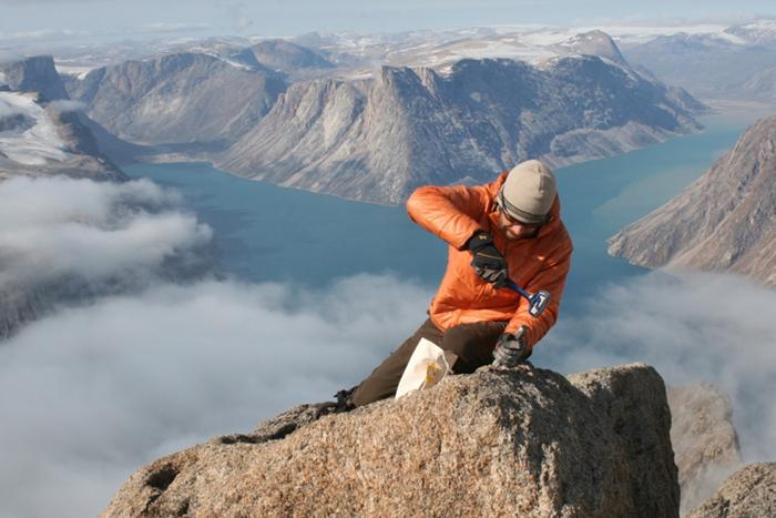 A man in an orange jacket uses a chisel on a rock at the summit of a mountain. A large lake is in the background behind him.