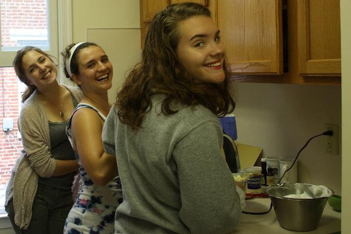 Three students smile at the camera as they pause from preparing cake batter.
