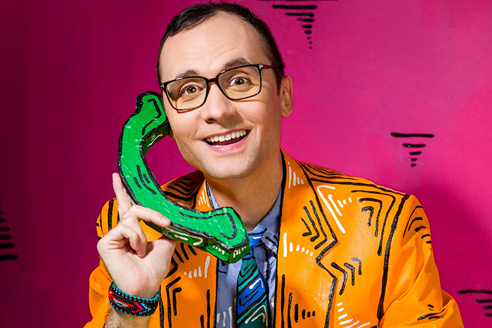 Comedia Joey Clift wearing an orange suit jacket and holding a green telephone.