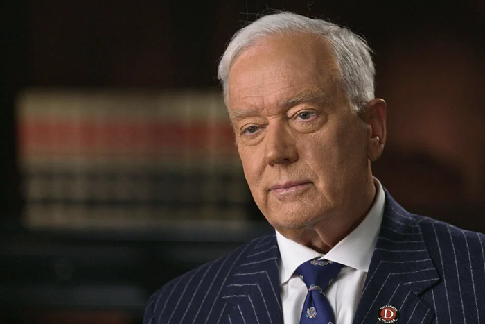 Screenshot of a man in a blue pinstripe suit in front of a bookcase.