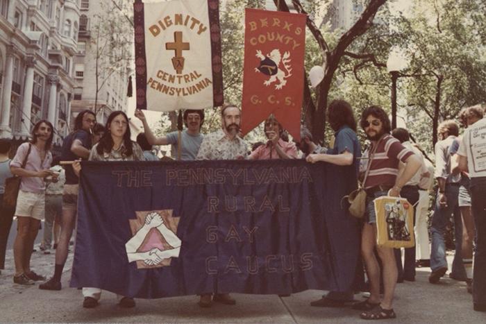Photo depicts members of the Pennsylvania Rural Gay Caucus in 1976 holding a purple banner that says "Pennsylvania Rural Gay Caucus" in a pride parade.