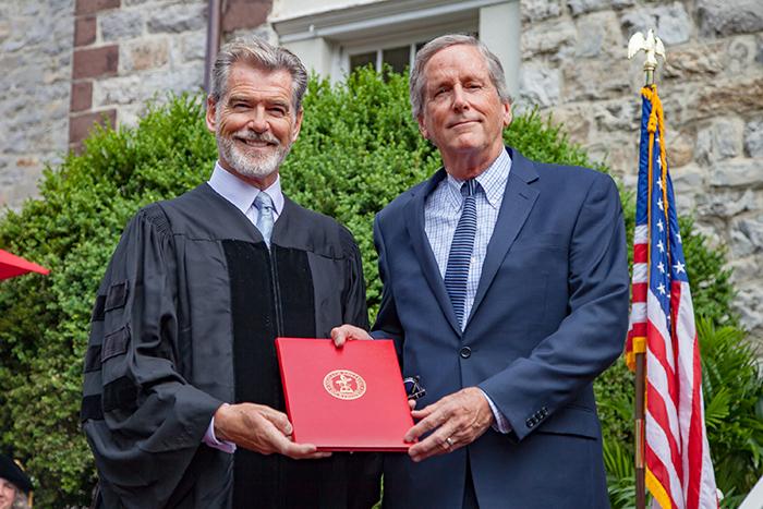 Pierce Brosnan stands next to Joel Reynolds. Both men are holding a red folder between them that contains the Rose-Walters Prize for Environmental Activism Citation