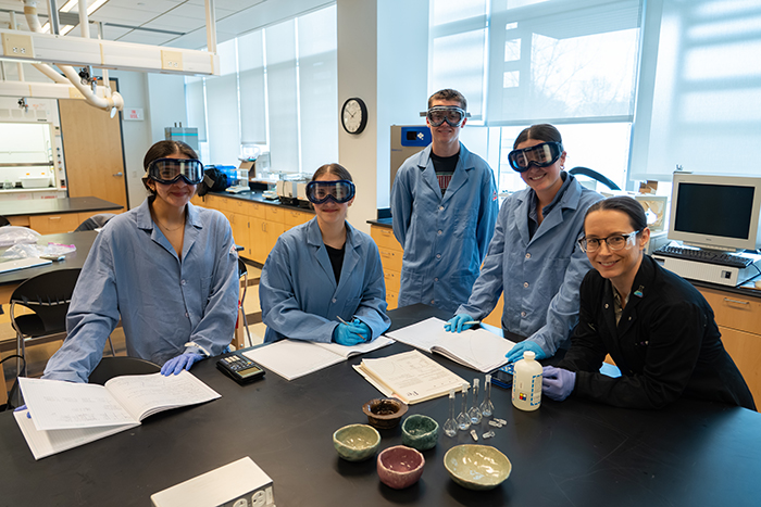 College students in lab coats and their professor gather around a table in the lab.