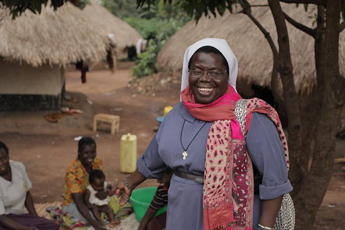 Portrait of Sister Rosemary Nyirumbe, wearing a blue top and a pink scarf, standing in an African village.