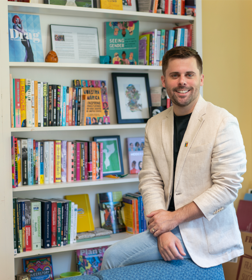 Photo of Tommy Walcott-Lee in a white blazer sitting in front of a bookshelf in his office. 