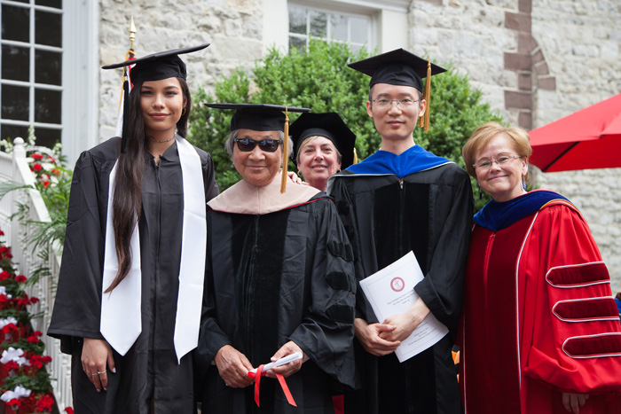 Adrian Zecha ’52, who was was presented Doctor of International Business honorary degree, poses with his daughter, Adya Zecha; 
Assistant Professor of International Business & Management Xiaolu Wang; and Ensign.