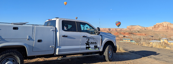 A truck drives across a rugged Western landscape. There are hot-air balloons in the distance.