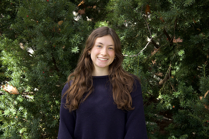 a young woman smiles at the camera while posing against a leafy backdrop.