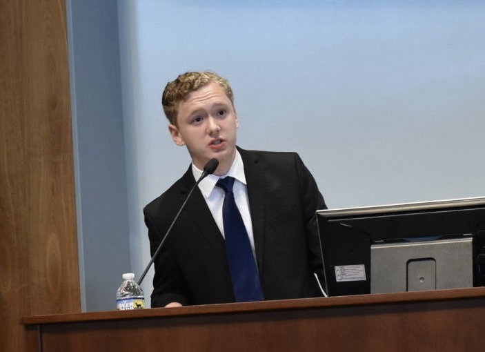 A young man in a suit speaks at a podium